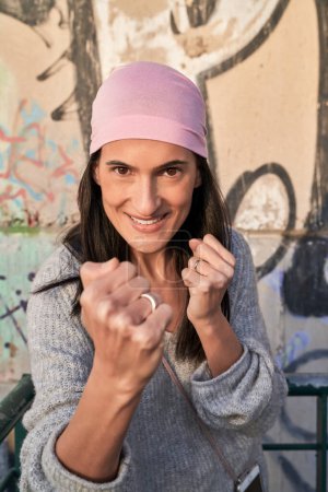 Photo for Young ethnic female in pink scarf showing fists and gesture of defense against breast cancer near wall with graffiti - Royalty Free Image