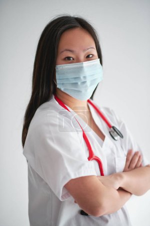 Foto de Vista lateral de mujer confiada en uniforme médico con estetoscopio cruzando brazos y mirando a la cámara sobre fondo blanco - Imagen libre de derechos