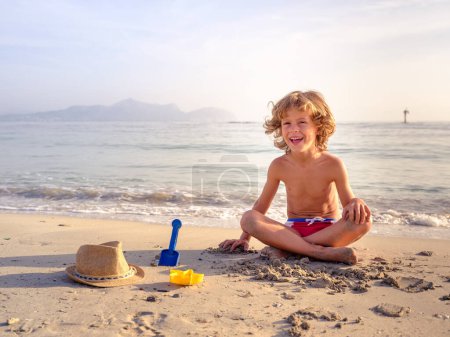 Photo for Full body of cheerful curly haired boy sitting with crossed legs on sandy seashore and playing with plastic toys in sunny day - Royalty Free Image