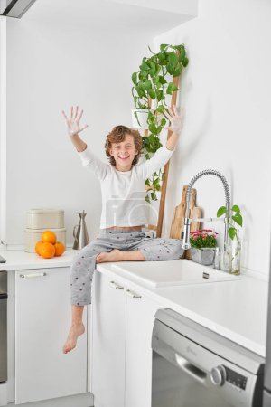 Photo for Full body of happy barefoot curly haired boy in pajama raising arms up while sitting on counter near sink and doing morning hygiene procedure - Royalty Free Image