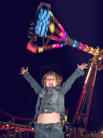 Photo for Amazed boy looking at camera with raised arms while standing near flowing ranger attraction in amusement park in evening time - Royalty Free Image