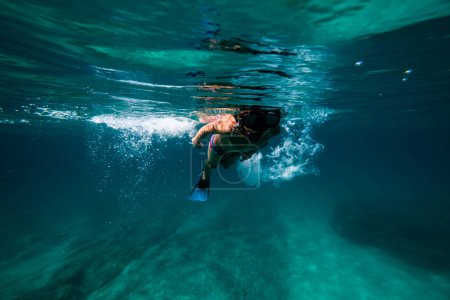 Téléchargez les photos : De dessous du corps complet préadolescent garçon en masque de plongée et nageoires nageant sous l'eau tout en observant le fond de la mer - en image libre de droit