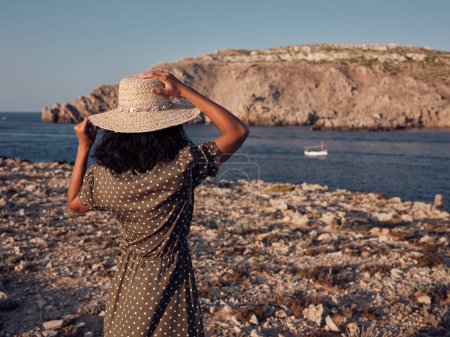 Photo for Back view of female wearing hat and dotted dress standing on rocky coast and observing boat floating in ocean on summer day - Royalty Free Image