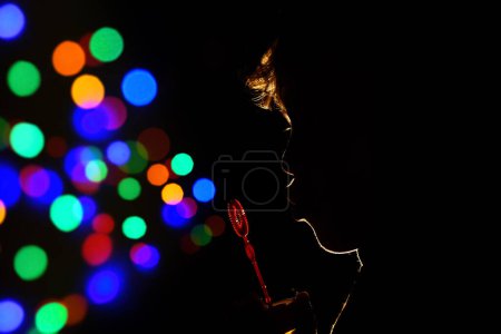 Photo for Side view silhouette of unrecognizable playful kid blowing soap bubbles while standing in dark room with round colorful Bokeh lights - Royalty Free Image