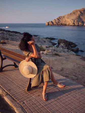 Foto de Mujer con ropa elegante sentada en el banco cerca del océano y disfrutando de las vacaciones de verano al atardecer mientras se apoya en la mano y mira hacia otro lado - Imagen libre de derechos