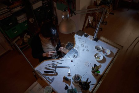 Téléchargez les photos : Angle élevé du joaillier féminin anonyme aplatissant le métal avec la lime à main assise au bureau avec la lampe et divers outils dans l'atelier sombre - en image libre de droit