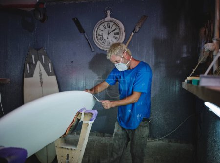 Photo for Side view of adult blond haired master in respirator mask grinding white surfboard placed in shaping racks while working in workshop - Royalty Free Image