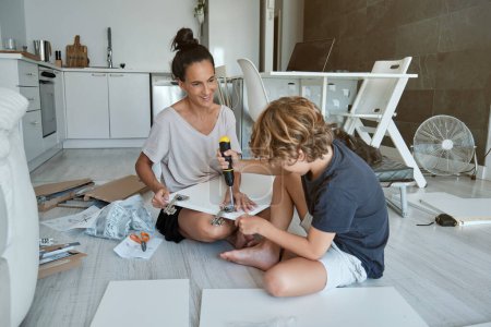 Photo for Full body of positive woman and boy in domestic clothes sitting on floor near instruction and screwing hinges to wooden door while assembling furniture in light kitchen - Royalty Free Image