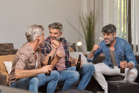 Téléchargez les photos : Fans masculins matures excités avec des bouteilles de bière s'amuser ensemble et célébrer la victoire pendant le match dans le salon à la maison - en image libre de droit