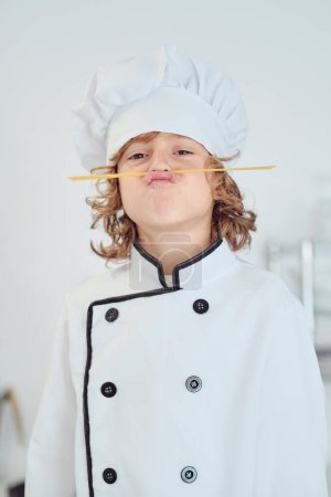 Téléchargez les photos : Adorable garçon préadolescent en costume de chef grimaçant et imitant la moustache avec des bâtons de spaghetti et regardant la caméra - en image libre de droit