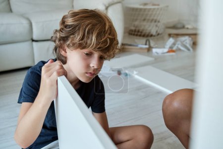 Photo for Concentrated curly haired preteen child sitting on floor and helping crop mother to assemble furniture in living room - Royalty Free Image