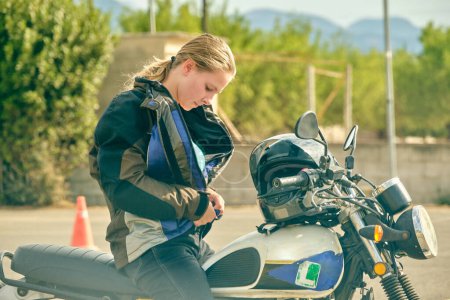 Photo for Side view of serious female motorcyclist sitting on contemporary motorbike and putting on jacket on motordrome - Royalty Free Image