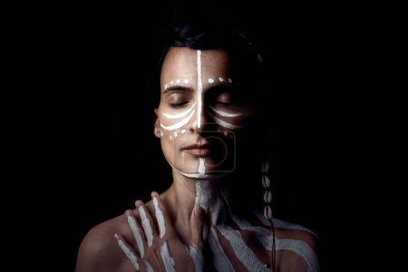 Téléchargez les photos : Portrait de jeune mannequin amérindienne avec body art et les yeux fermés touchant la poitrine sur fond noir en studio - en image libre de droit