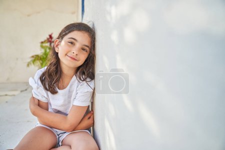 Photo for Cute smiling preteen girl with long brown hair in casual clothes sitting on sidewalk with crossed arms and leaning on concrete wall while looking at camera - Royalty Free Image