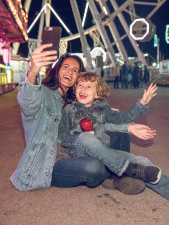 Téléchargez les photos : Femme positive avec un enfant assis sur le sol et prenant autoportrait sur téléphone portable tout en passant la nuit dans un parc d'attractions - en image libre de droit