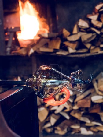 Photo for Shiny glass vase with metal tube placed near burning fire and pile of firewood in workshop - Royalty Free Image