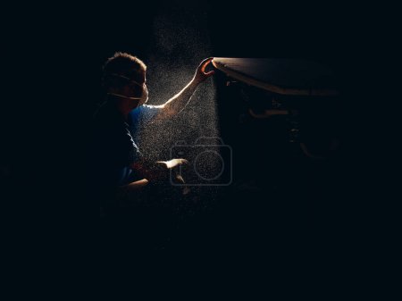 Photo for Side view of male master in respirator sitting on haunches in dark dusty garage and making surfboard under sunlight - Royalty Free Image