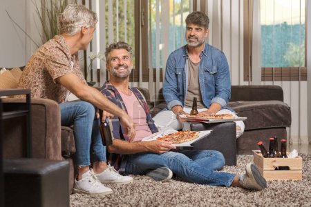Photo for Content adult men in casual clothes smiling and looking at each other while eating pizza and drinking beer in cozy living room - Royalty Free Image