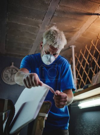 Photo for From below of focused male shaper in respiratory mask polishing tail of surfboard with abrasive fabric in workshop - Royalty Free Image