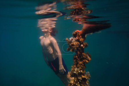 Téléchargez les photos : Vue latérale du garçon anonyme en short nageant jusqu'à reprendre son souffle près du corail se reflétant dans l'eau le jour de l'été pendant les vacances - en image libre de droit