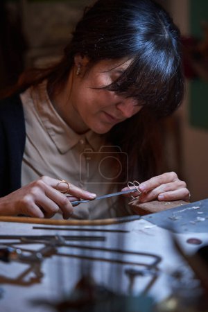 Téléchargez les photos : Orfèvre féminin ethnique adulte concentré aux cheveux foncés aplatissant les bords des bijoux avec une lime plate tout en travaillant dans un atelier traditionnel - en image libre de droit