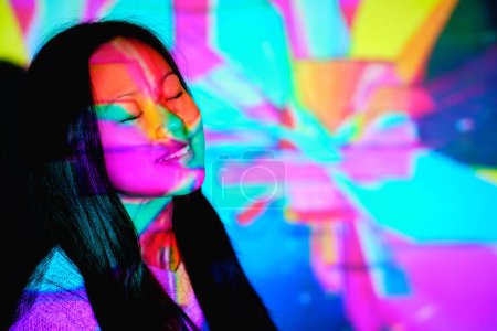 Téléchargez les photos : Contenu Femme asiatique avec les yeux fermés debout sous des lumières lumineuses sur le visage près du mur multicolore en studio avec un éclairage lumineux - en image libre de droit