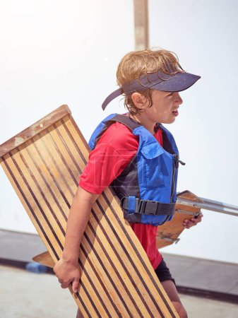 Photo for Side view of little boy in casual clothes and cap with wooden bench looking away on sunny day - Royalty Free Image