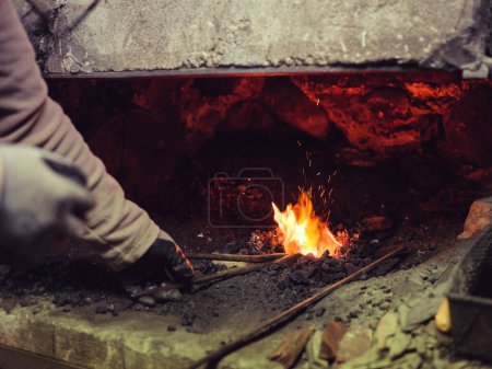 Téléchargez les photos : Cultiver faussaire masculin méconnaissable dans des gants de protection debout pinces et brûler du métal dans le four tout en travaillant dans un atelier de forge - en image libre de droit