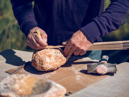 Photo for Crop unrecognizable mature craftsman making marks on solid rough stone using ruler and pencil while creating rock craft on workbench in sunny day - Royalty Free Image