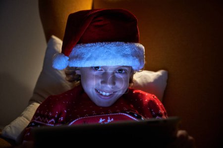 Téléchargez les photos : Adorable petit garçon dans le chapeau de Père Noël et pull chaud en utilisant la tablette tout en étant assis dans un fauteuil doux et en regardant la caméra - en image libre de droit