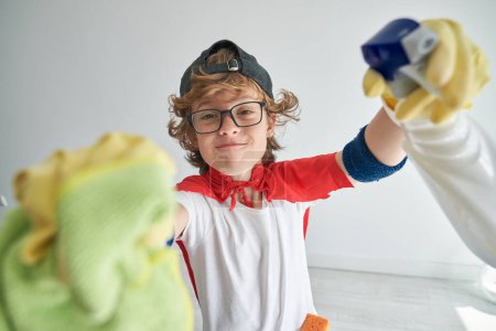 Foto de Niño satisfecho con anteojos y ropa de superhéroe limpiando cámara con paño y pulverizador en estudio de luz - Imagen libre de derechos
