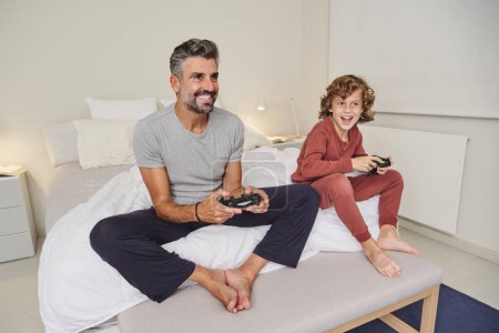 Foto de Longitud completa de sonreír descalzo maduro padre y niño jugando videojuego en el dormitorio - Imagen libre de derechos