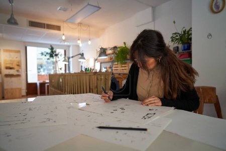 Téléchargez les photos : Artiste féminine occupée dessinant des croquis sur des feuilles de papier tout en créant des croquis d'accessoires de bijoux faits à la main en studio - en image libre de droit