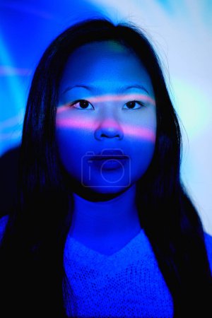Téléchargez les photos : Belle femelle asiatique avec de longs cheveux noirs regardant vers l'avant avec un visage rêveur tout en se tenant dans une pièce sombre avec des lumières roses et bleues éclatantes - en image libre de droit