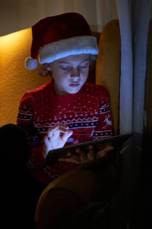 Foto de Adorable niño pequeño en Santa sombrero sentado en sillón en habitación oscura y mirando hacia abajo mientras navega tableta - Imagen libre de derechos