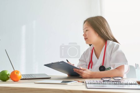 Photo for Side view of concentrated Asian female doctor in uniform writing on clipboard while working on laptop at table in clinic - Royalty Free Image