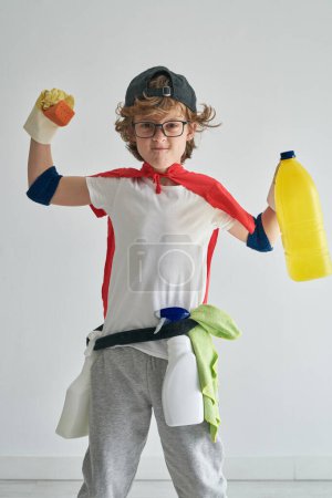 Photo for Positive preteen boy smiling and looking at camera while standing in red superhero cape on shoulders and showing soap and detergent in bottle against gray wall - Royalty Free Image
