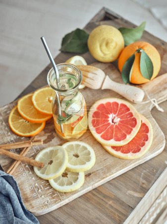 Foto de Botella de sabrosa limonada servida con varios cítricos en rodajas y palitos de canela sobre tabla de cortar con limón entero y naranja sobre mesa de madera - Imagen libre de derechos