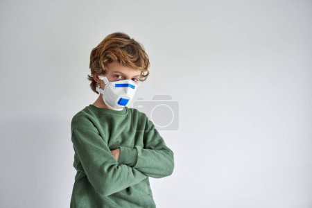 Photo for Preteen boy in medical respirator with arms crossed during quarantine restrictions during covid19 pandemic and looking at camera on gray background - Royalty Free Image