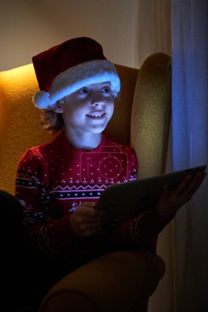 Foto de Encantador niño en Santa sombrero y suéter usando tableta mientras está sentado en un cómodo sillón y mirando hacia otro lado - Imagen libre de derechos