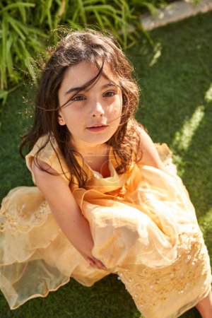 Téléchargez les photos : D'en haut de l'enfant préadolescent calme en élégante robe jaune assis sur l'herbe dans le jardin et regardant la caméra - en image libre de droit