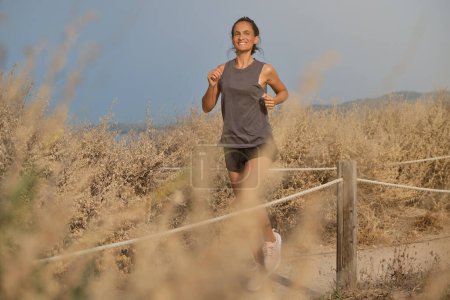 Téléchargez les photos : Pleine longueur d'athlète féminine positive en vêtements de sport souriant et regardant loin tout en faisant du jogging sur une promenade sablonneuse dans la campagne - en image libre de droit