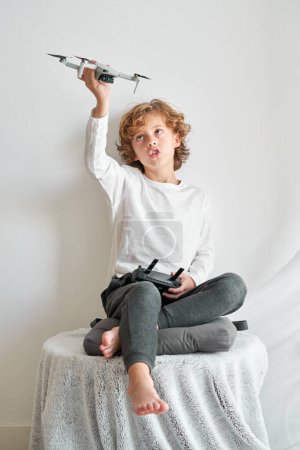 Photo for Full length of blond kid sitting on table and looking up while having fun with toy drone - Royalty Free Image