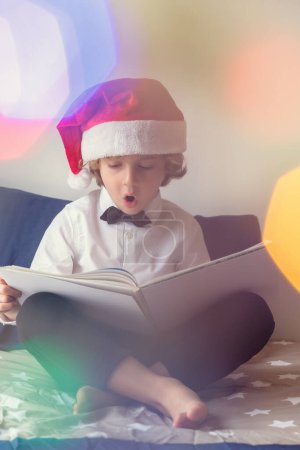 Foto de Cuerpo completo de chico interesado en ropa formal y libro de lectura de sombrero de Santa con la boca abierta mientras está sentado en la cama durante las vacaciones de Navidad - Imagen libre de derechos