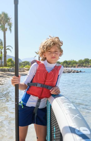 Téléchargez les photos : Enfant sérieux avec pagaie et planche SUP en gilet de sauvetage regardant la caméra tout en se tenant debout dans la mer bleue ondulante le jour d'été lumineux - en image libre de droit
