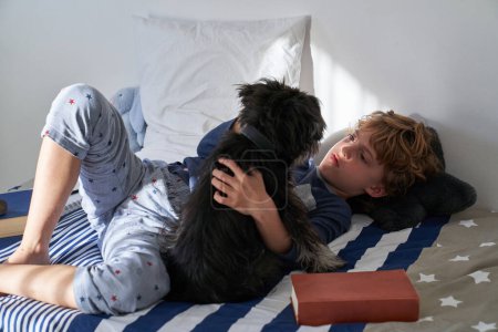 Foto de Sereno chico de pelo rizado relajándose en la cama mientras acaricia cachorro esponjoso en el dormitorio de luz en casa - Imagen libre de derechos