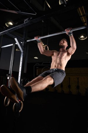 Photo for From below full body of focused muscular male doing pull ups on bar in gym on black background - Royalty Free Image