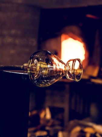 Foto de Jarrón de vidrio transparente con tubo de metal para soplado de vidrio colocado cerca del horno de combustión y leña en un taller profesional - Imagen libre de derechos