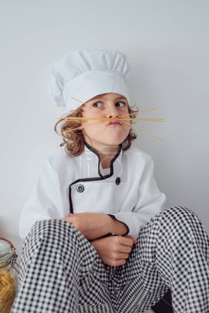 Téléchargez les photos : Adorable garçon préadolescent en costume de chef et casquette grimacant à l'aide de bâtonnets de spaghetti comme moustache imaginaire tout en étant assis sur le comptoir de la cuisine - en image libre de droit