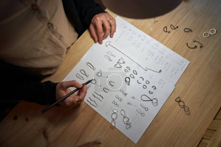 Foto de Vista superior de orfebre anónimo sin rostro dibujando bocetos de joyería con lápiz en hoja de papel sobre mesa de madera - Imagen libre de derechos